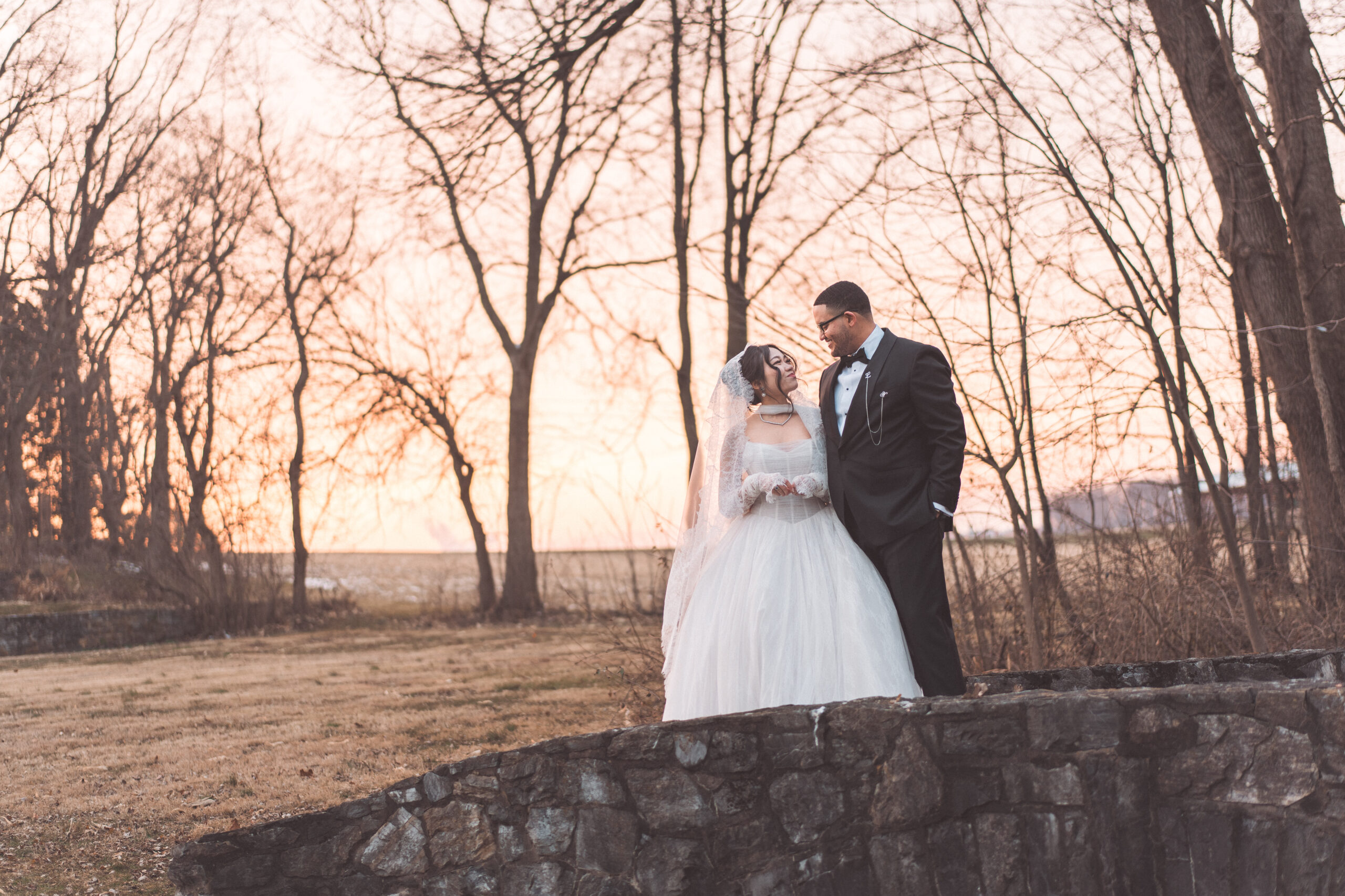 Newlyweds standing together outdoors at sunset with bare trees glowing in the warm evening light.