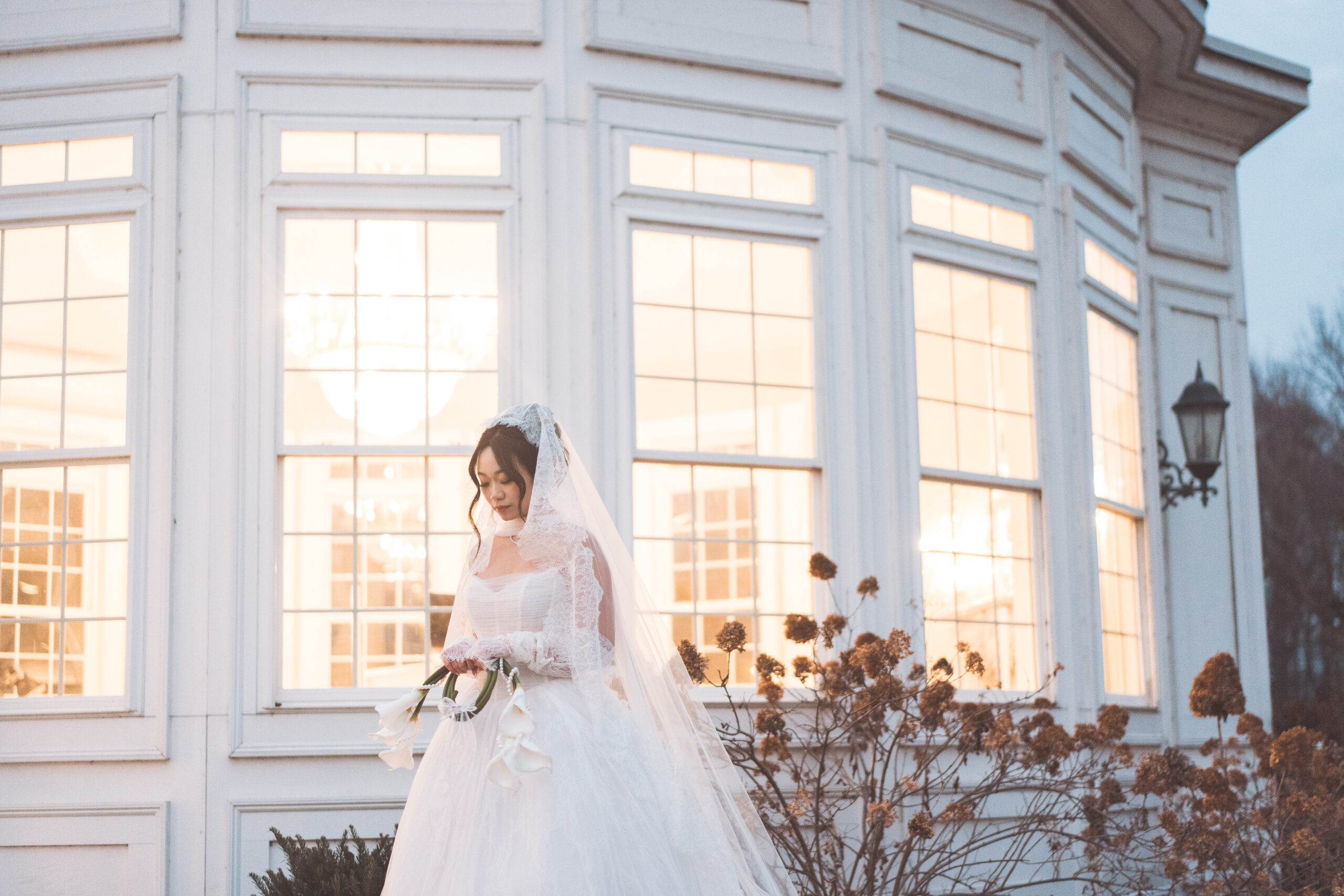 Bride holding a bouquet of white calla lilies outside the glowing windows of Cameron Estate Inn.