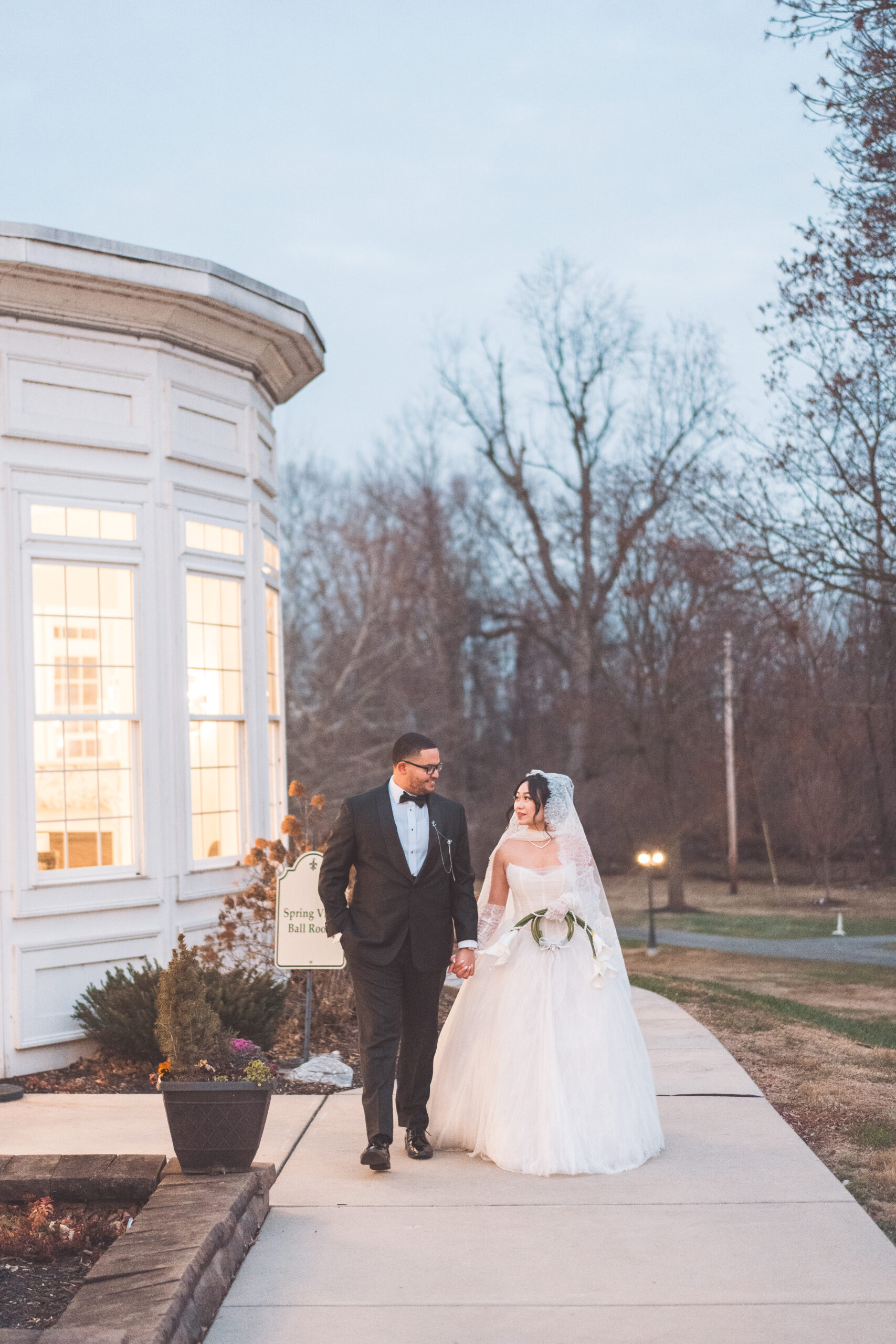 Bride and groom walking hand in hand outside the venue at dusk with the bride carrying white calla lilies.