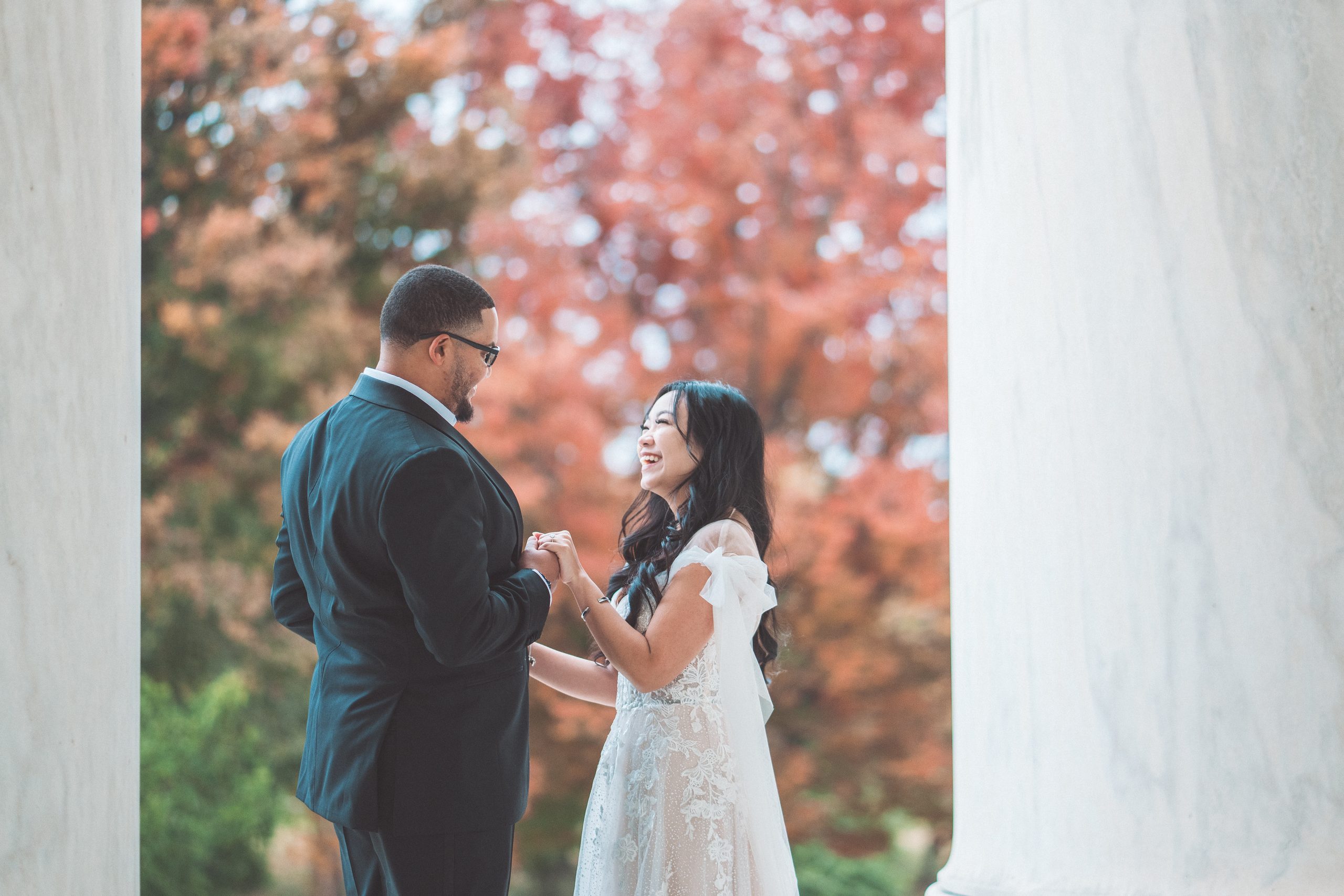 Couple laughing together between white marble columns with colorful fall foliage behind them during their Jefferson Memorial engagement photos.