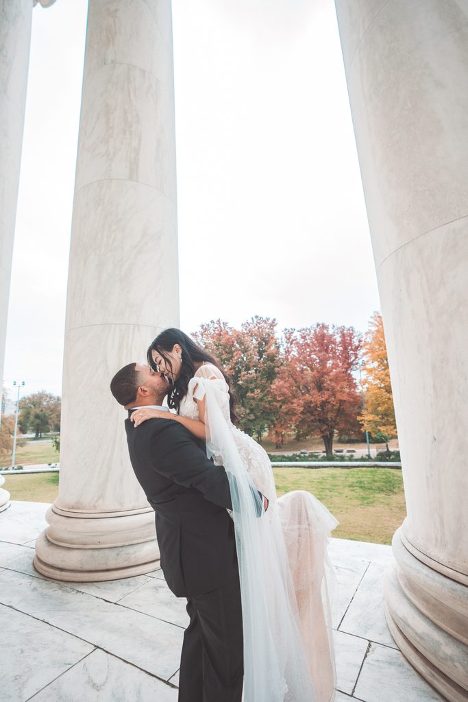 Groom-to-be lifting and kissing his fiancée between the marble columns during Jefferson Memorial engagement photos.