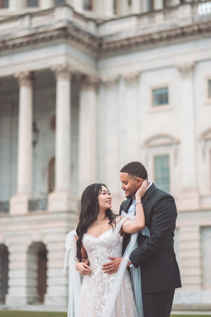 Engaged couple smiling and embracing in front of historic Washington DC architecture.