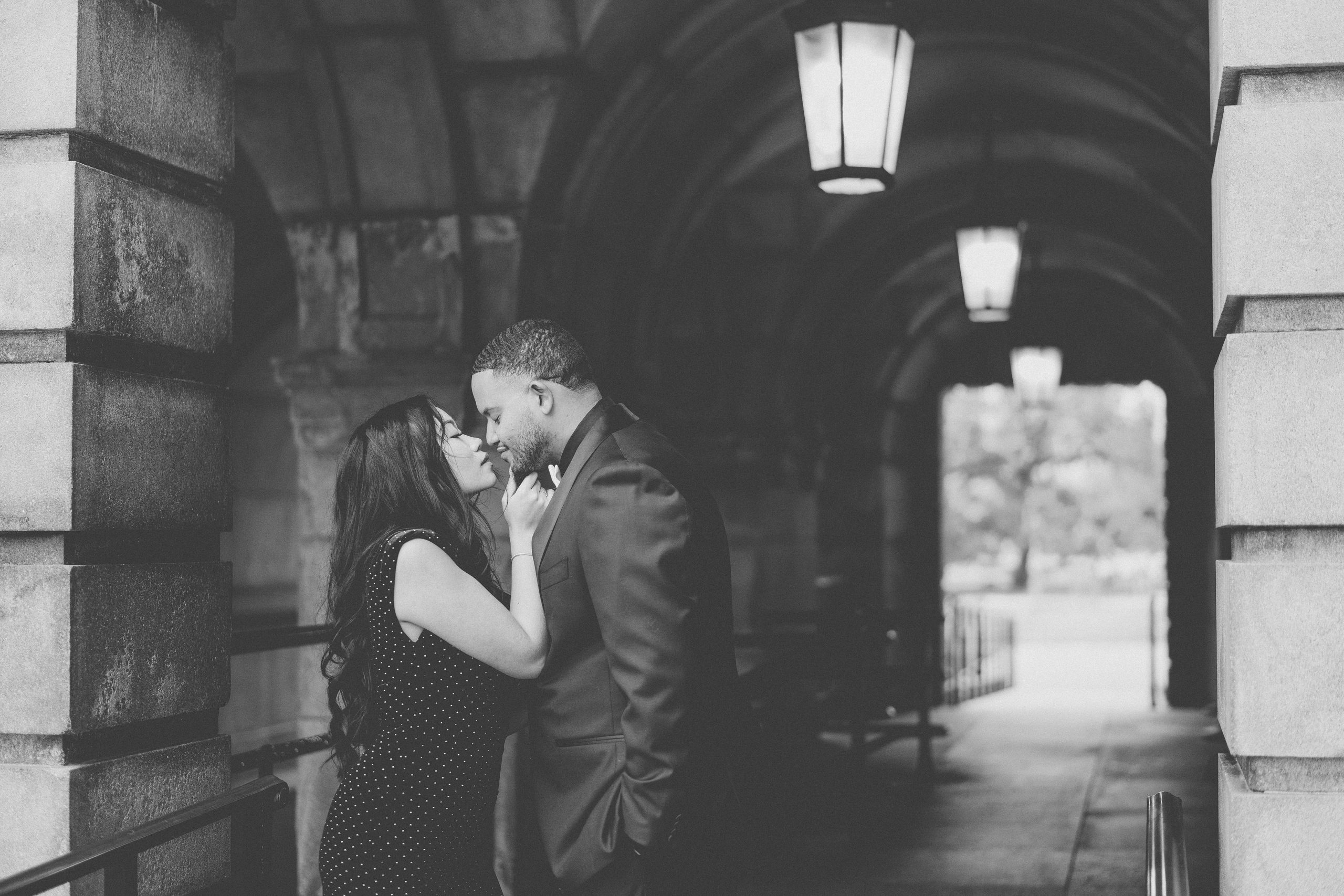 Black-and-white engagement photo of a couple sharing a kiss beside historic stone architecture.
