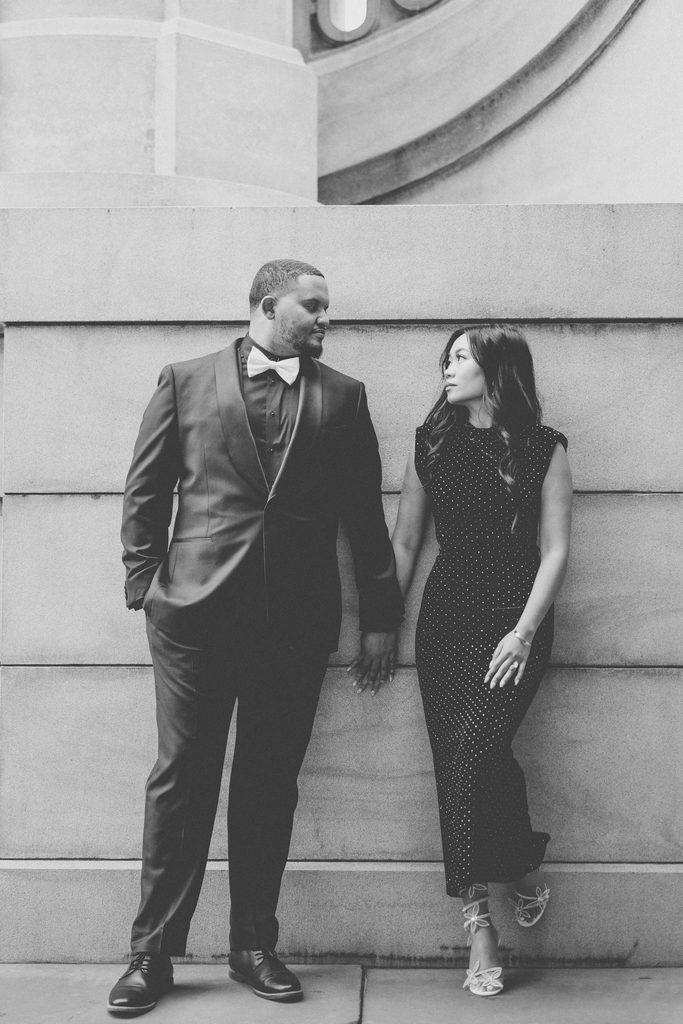 Black-and-white full-length portrait of a couple holding hands against a stone wall in Washington DC.