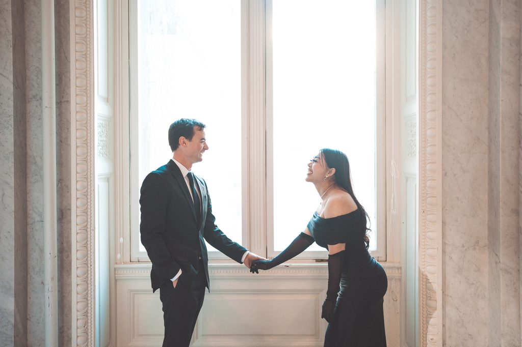 Candid Library of Congress Engagement Photos of a couple holding hands and laughing together beside a large window.