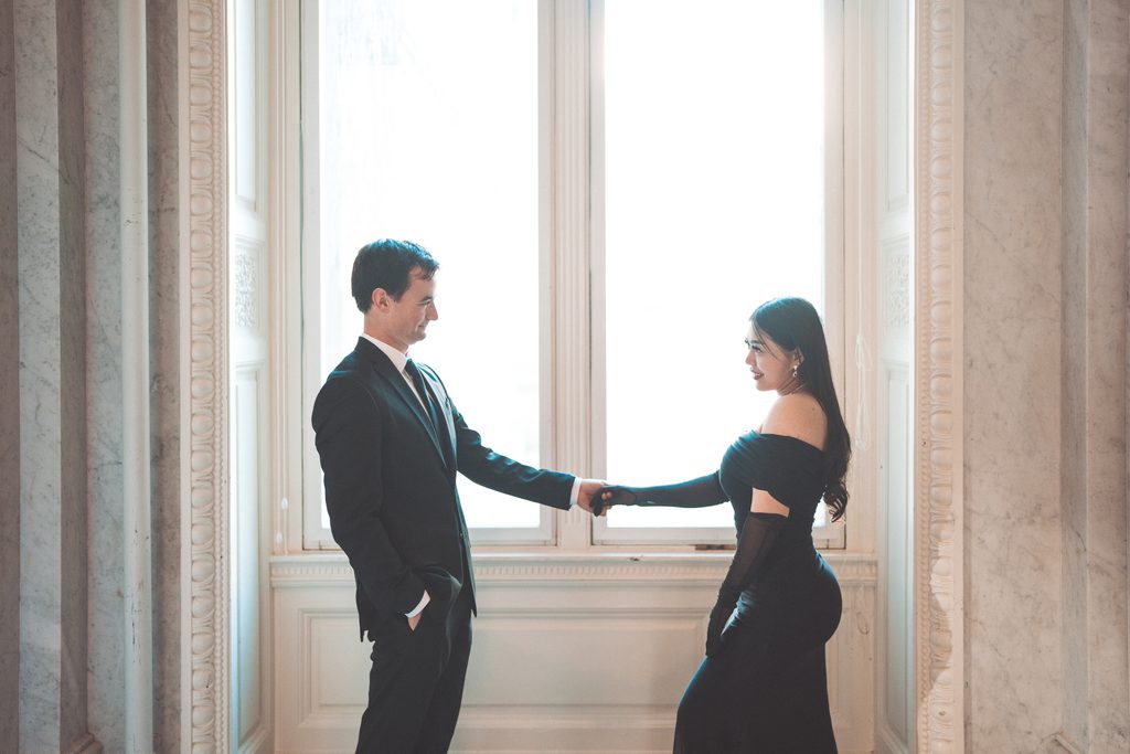 Couple holding hands and smiling at each other beside a bright window inside the Library of Congress.