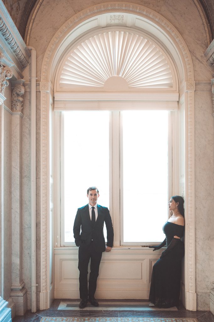 Couple posing across from each other beside a large window inside the Library of Congress.