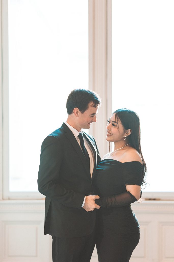 Romantic portrait of a couple smiling at each other beside a bright window inside the Library of Congress.