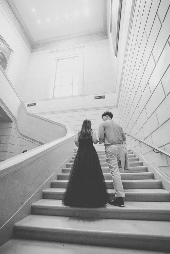 Black-and-white photo of a couple walking hand in hand up a sweeping marble museum staircase.