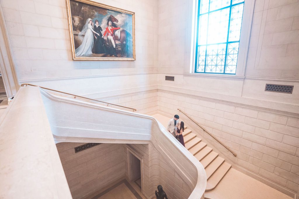 National Gallery of Art engagement photos of a couple walking together along a bright marble staircase beneath a framed painting.