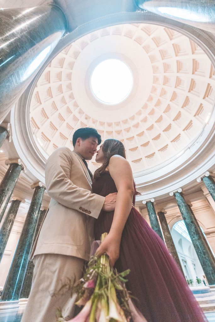 Couple leaning in for a kiss beneath the dramatic domed ceiling and marble columns inside the museum.