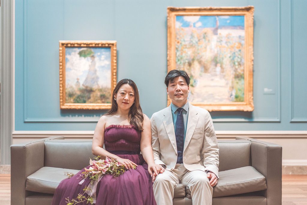 Couple sitting side by side on a museum bench with colorful framed paintings behind them.