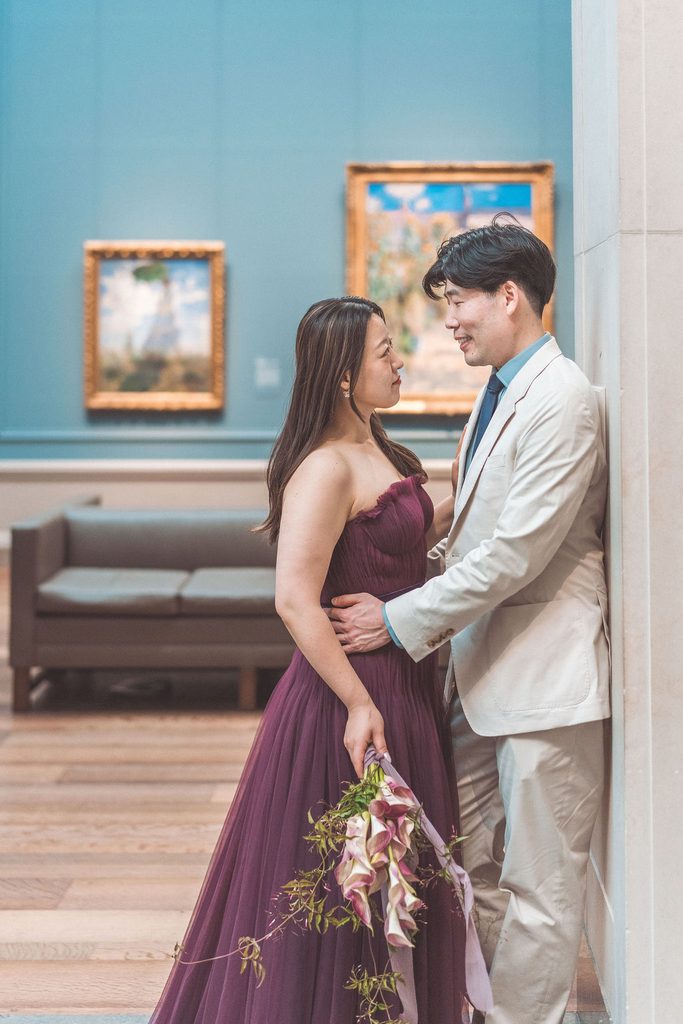 Couple smiling at each other in a blue-walled gallery, with the woman holding a cascading floral bouquet.