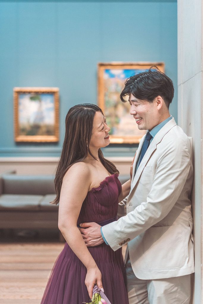 Couple smiling at each other in a blue-walled gallery surrounded by framed paintings.