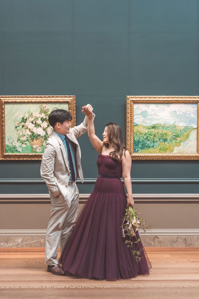 Couple playfully twirling in front of framed paintings in a blue-green gallery, with the woman holding a cascading bouquet.