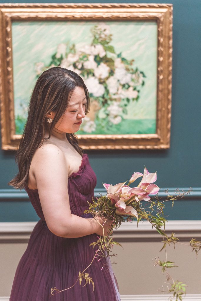 A bride looks down at her cascading bouquet with art on a blue wall behind her.