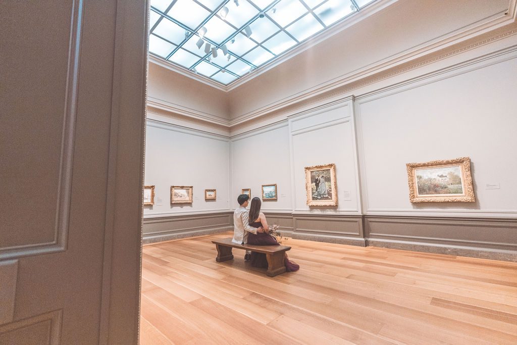 Couple sitting together on a wooden bench while looking at paintings in a bright gallery.