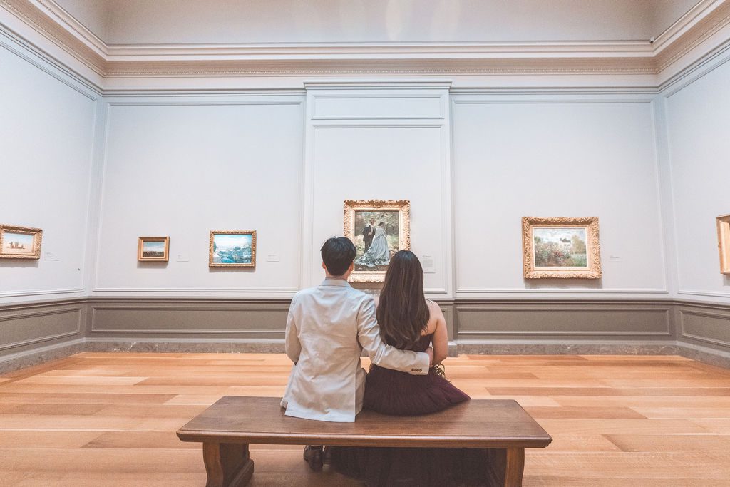 Couple sitting together on a wooden bench admiring paintings inside an elegant museum gallery.