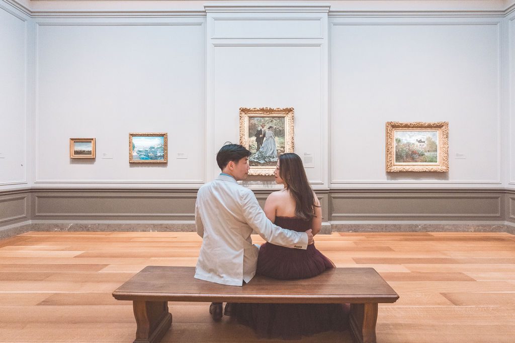 Couple sitting closely together on a wooden bench while viewing paintings in a bright museum gallery.