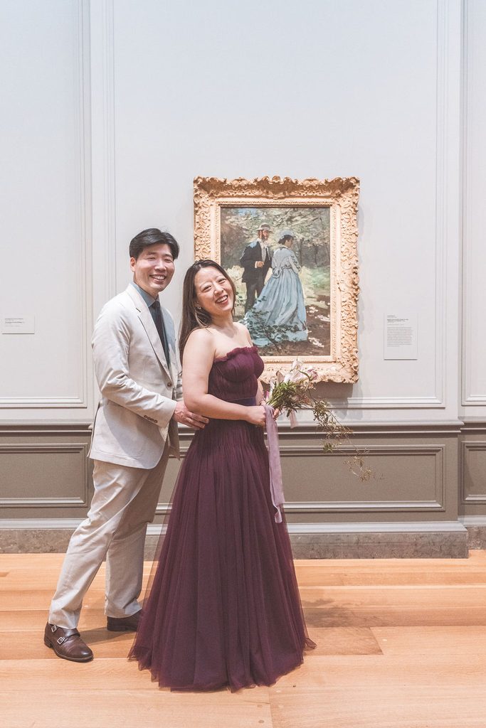 Couple laughing together in front of a framed painting, with the woman holding a cascading floral bouquet.