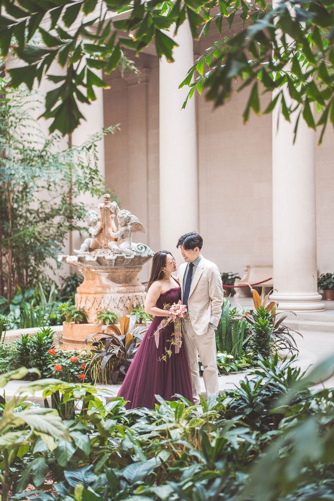 National Gallery of Art engagement photos of a couple standing together among lush greenery and a decorative fountain in the museum courtyard.