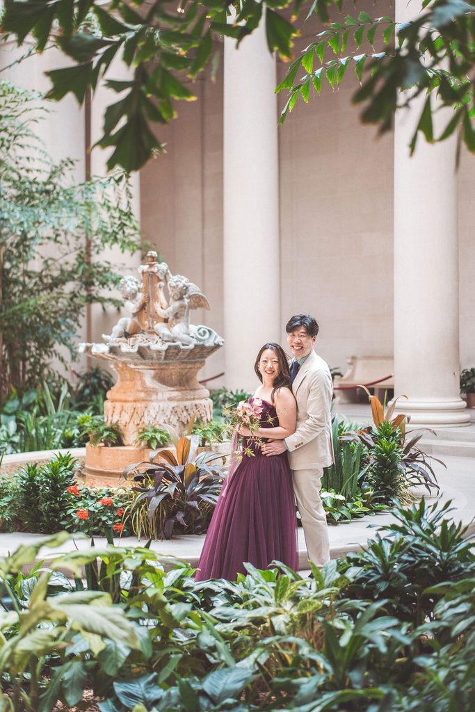 National Gallery of Art engagement photos of a couple laughing together among lush plants and a decorative courtyard fountain.