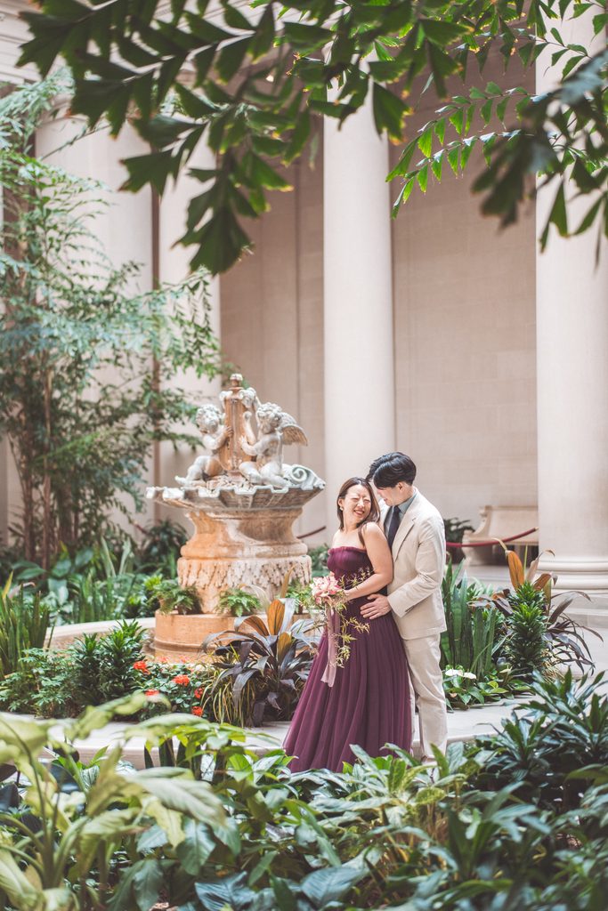 National Gallery of Art engagement photos of a couple embracing beside the fountain and lush greenery in the museum courtyard.
