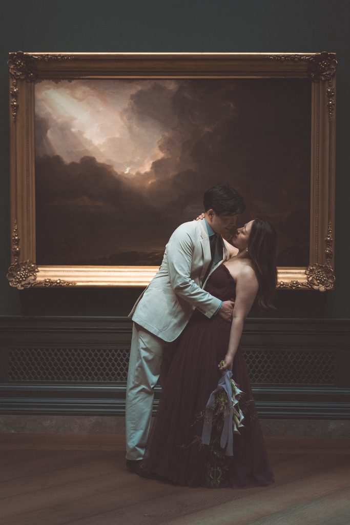 National Gallery of Art engagement photos of a couple sharing a romantic kiss beneath a dramatic landscape painting.
