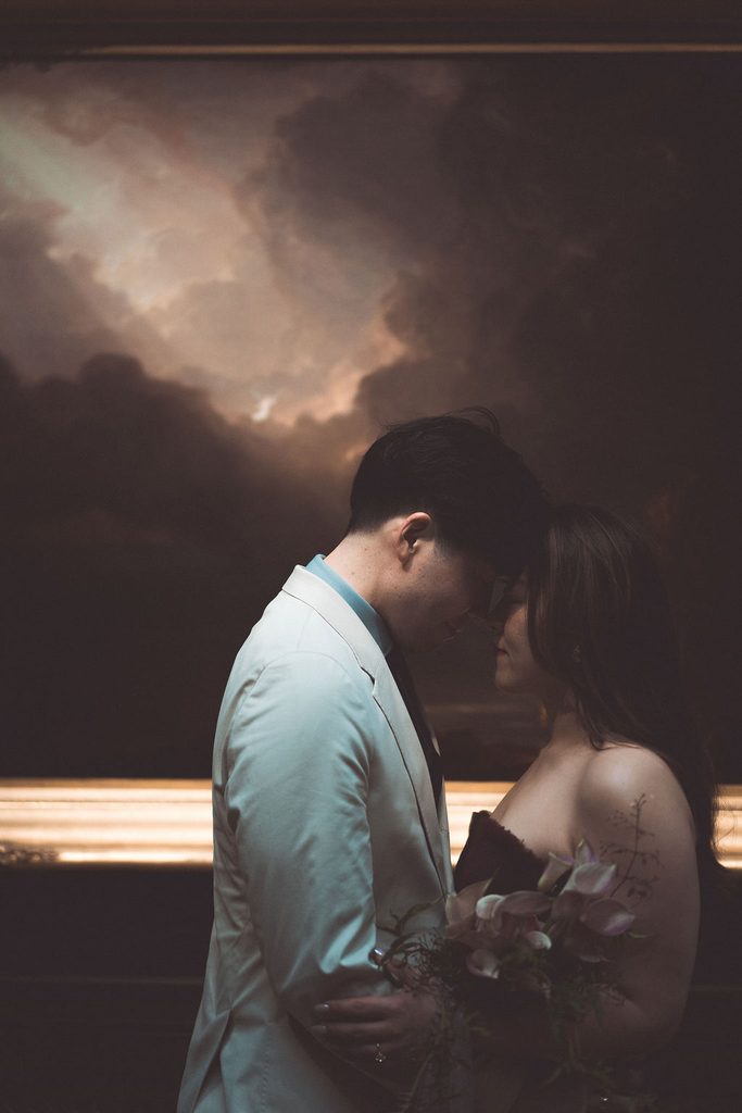 Couple sharing an intimate forehead-to-forehead moment in a dimly lit gallery with a floral bouquet in the foreground.