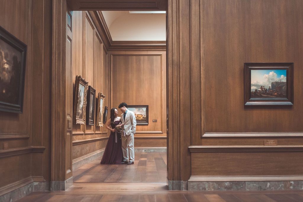National Gallery of Art engagement photos of a couple sharing a quiet moment at the end of a warm wood-paneled gallery lined with paintings.