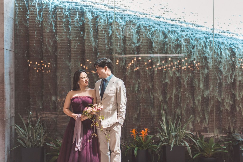 Couple smiling at each other beside the indoor water wall and greenery.
