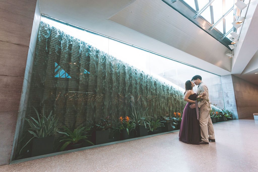 National Gallery of Art engagement photos of a couple sharing a quiet embrace beside a dramatic indoor water feature and greenery.