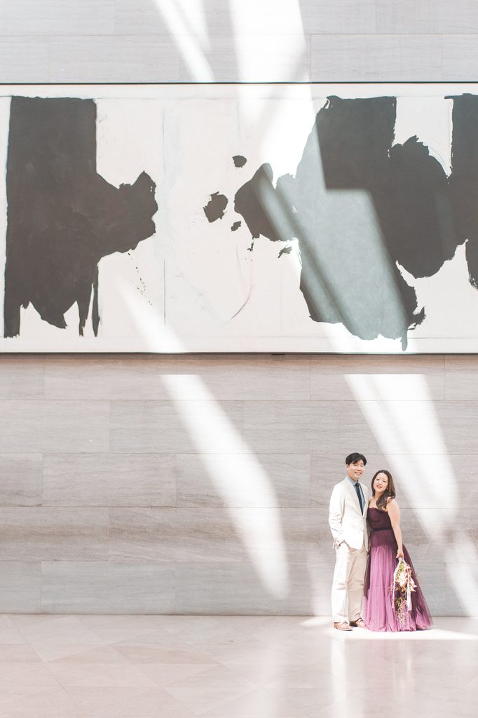 Couple standing together beneath a large abstract painting with dramatic natural light across the gallery wall.