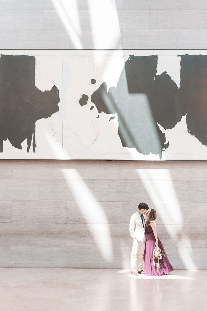 National Gallery of Art engagement photos of a couple kissing beneath dramatic beams of sunlight and a large abstract artwork.