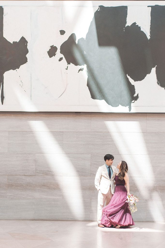Couple standing together beneath dramatic beams of sunlight against a minimalist marble wall.