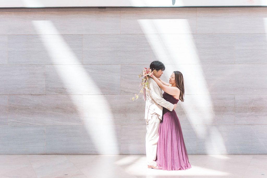 Couple embracing in a beam of natural light against a minimalist museum wall.