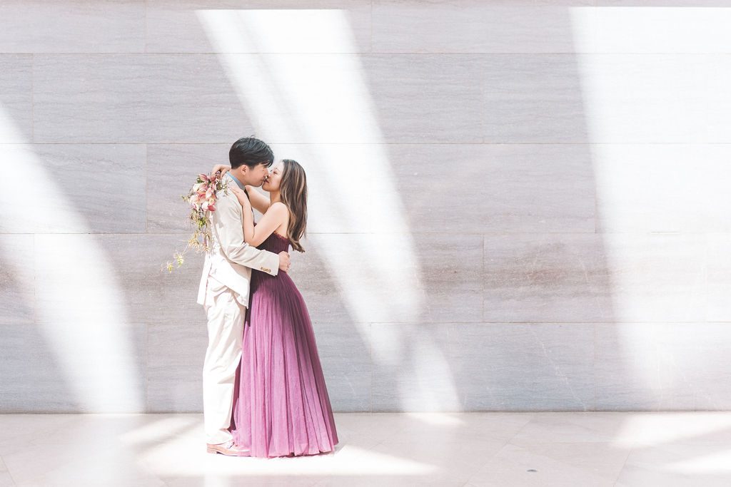 National Gallery of Art engagement photos of a couple kissing beneath dramatic natural light against a marble gallery wall.